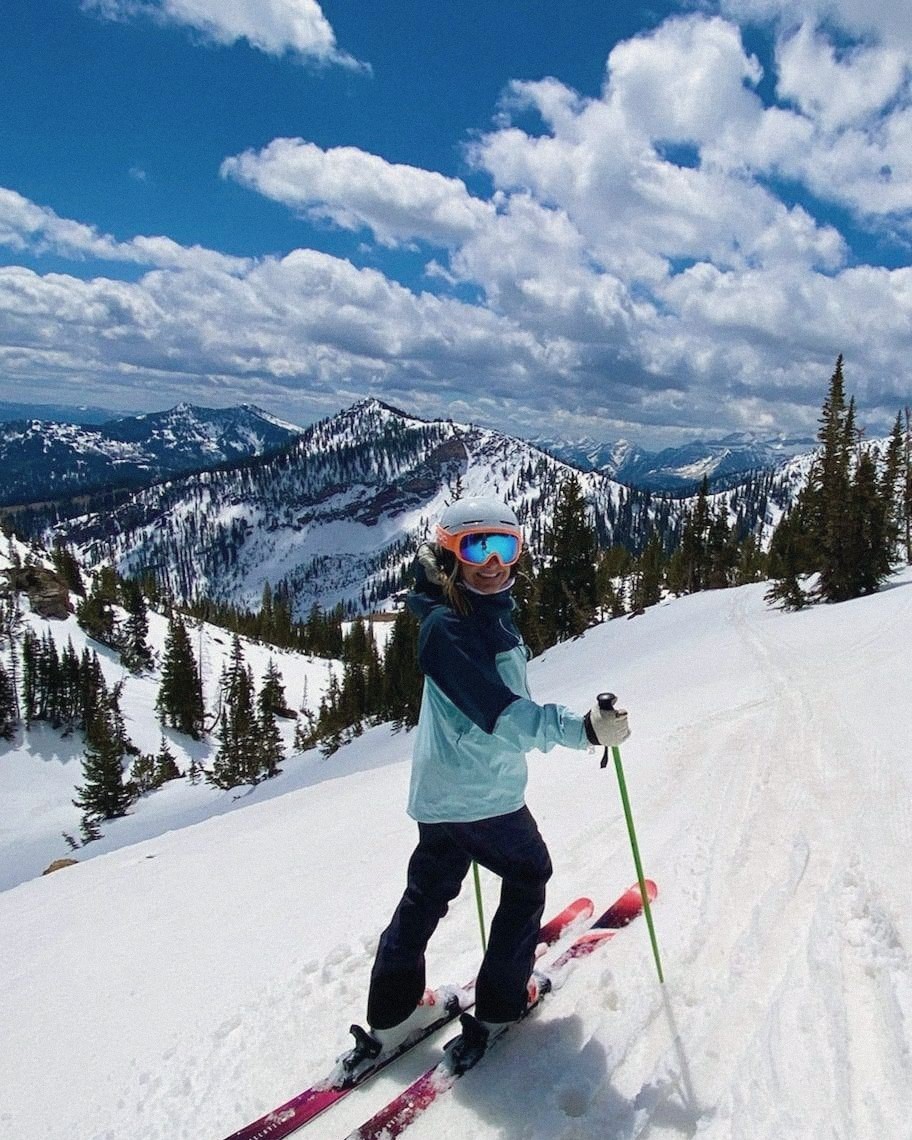 Sue skiing at Snowbird in a teal jacket and orange goggles, smiling back at the camera, with snowy peaks behind her