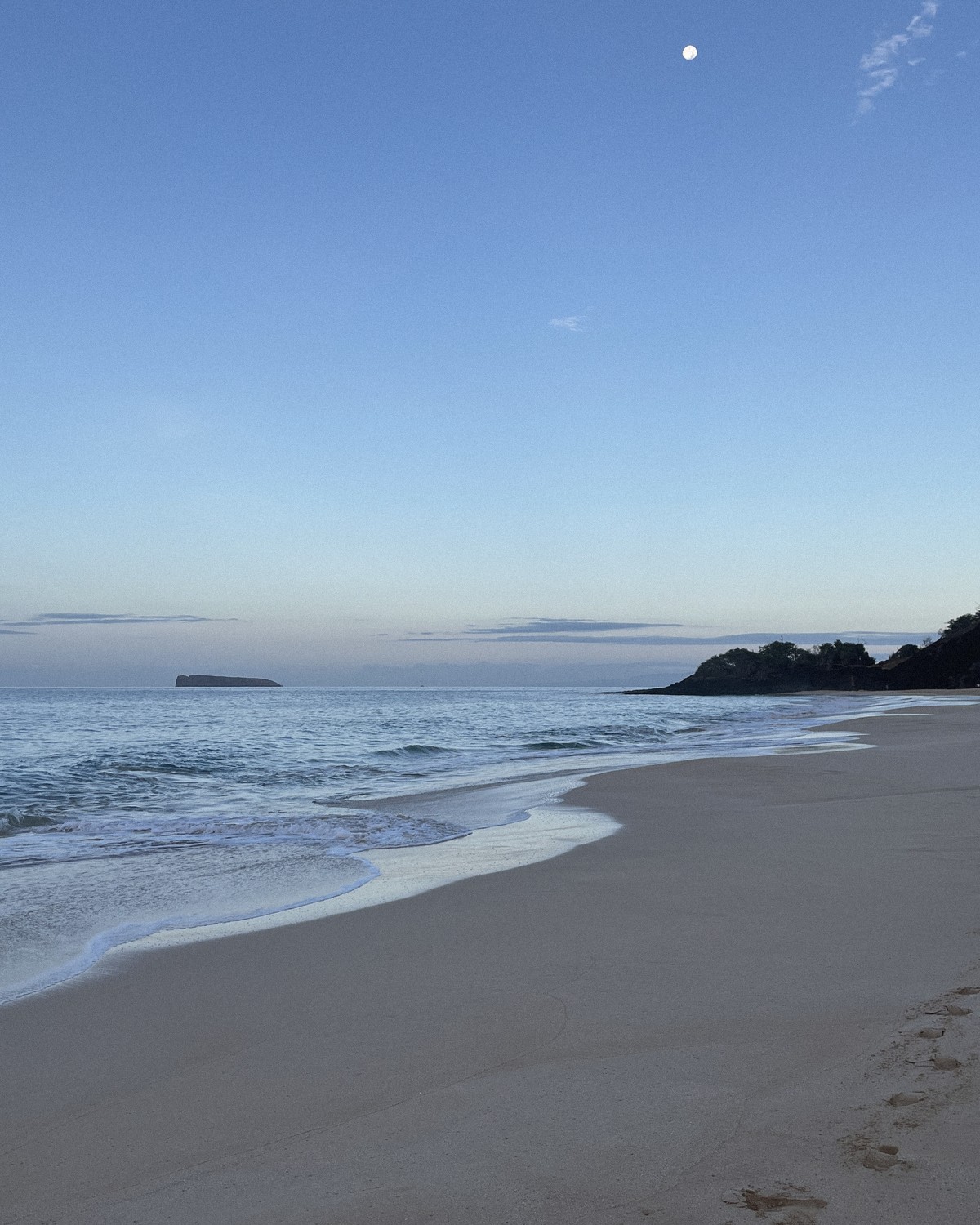 Big Beach, Maui, at first light: a long empty beach, gentle surf, the moon high in a pale blue sky and Molokini on the horizon
