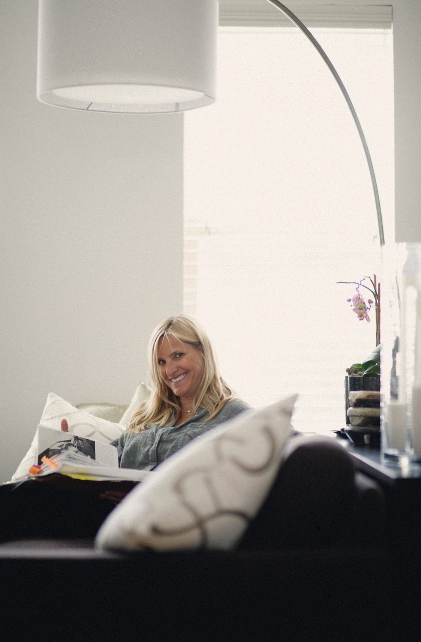 Sue Randall, smiling, sitting on a couch at home with a magazine, beside an arc lamp